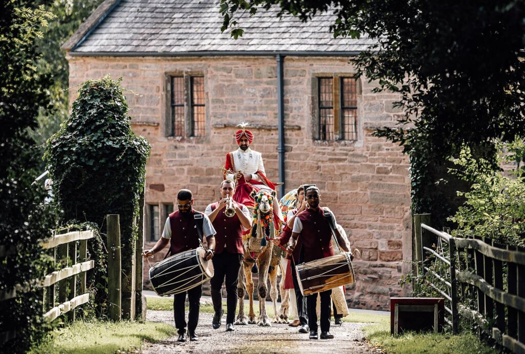 Wedding Photography Somerset Groom riding Camel to meet his bride at crowcombe court wedding venue in somerset, there is also with music drummers in front as they walk the long drive.