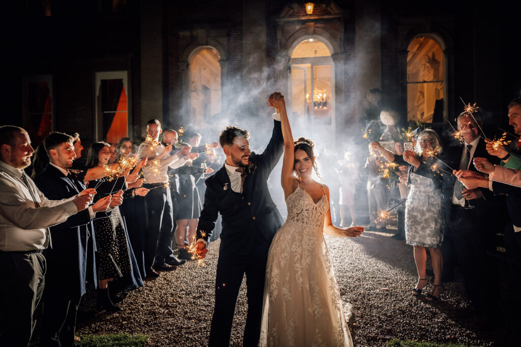 Bride and groom standing in front of crowcombe court laughing with sparklers in had enjoying a moment with all their guests at their fun wedding.