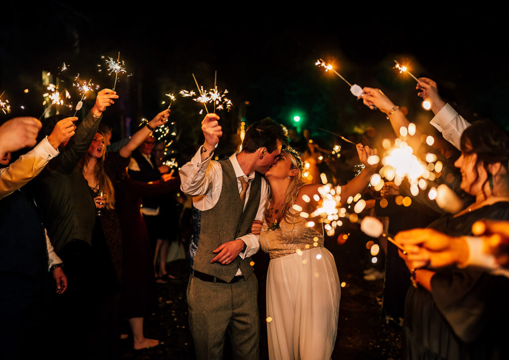 Bride and groom walking down sparkler line having a kiss while holding sparklers, surrounded by friends and family at their wedding at fairy hill, swansea.