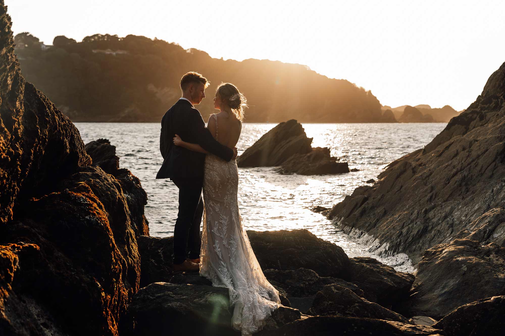 Bride and groom looking at each other while satnding on rocks overlooking sandy cove