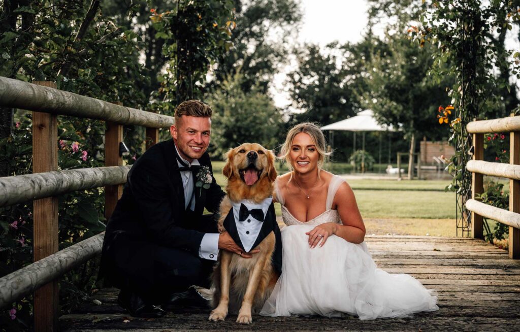 Bride and groom kneeling down with their golden retriever sat in between them at waterside country barn