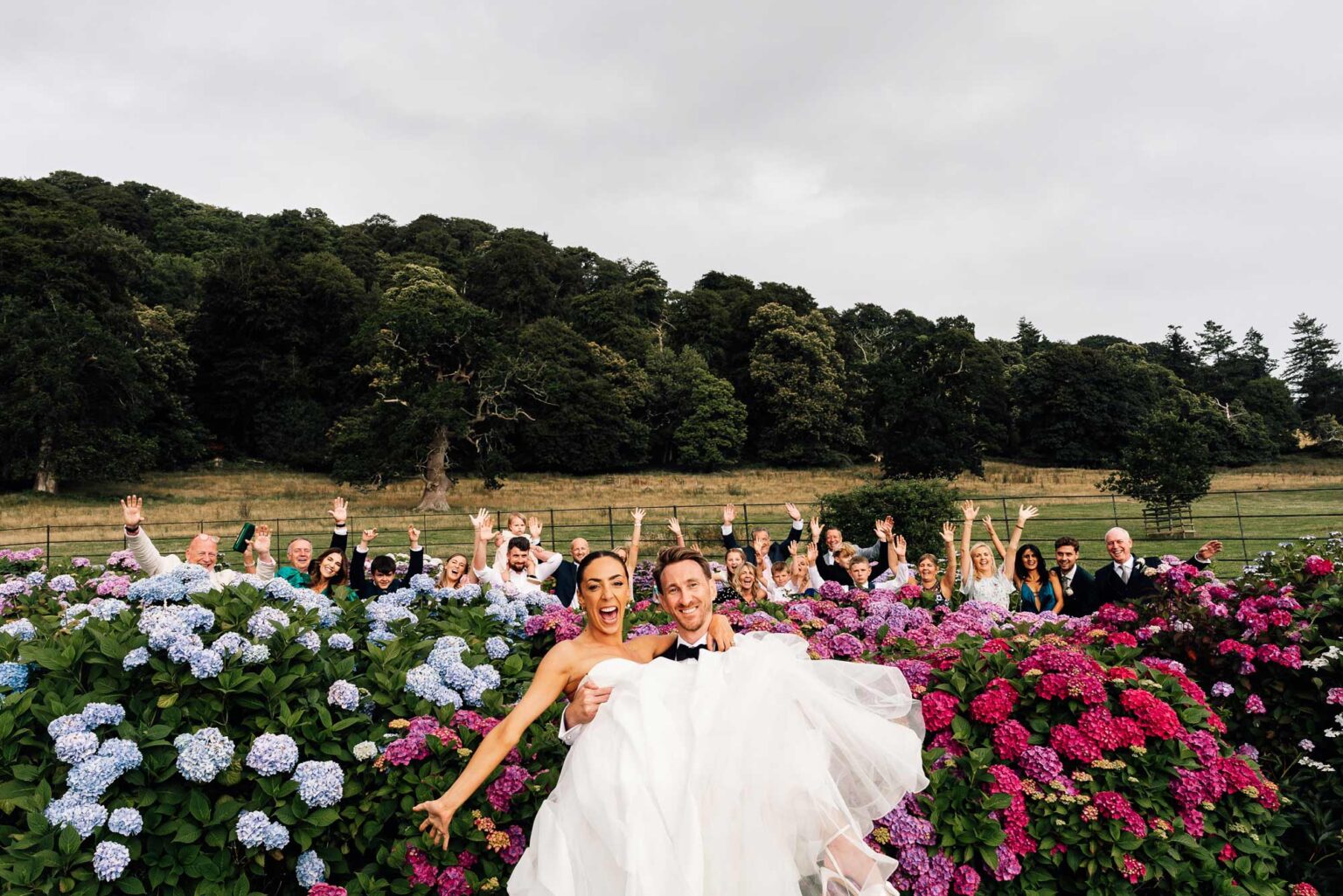 groom carrying bride in front of flower bed while his bridal party jump in the air at their wedding- Crowcombe court wedding Paul aston Photography Somerset wedding photographer