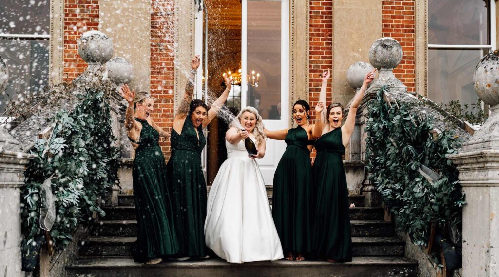 Bride and her bridesmaids on the steps of crowcombe celebrating with spraying a bottle of champagne.